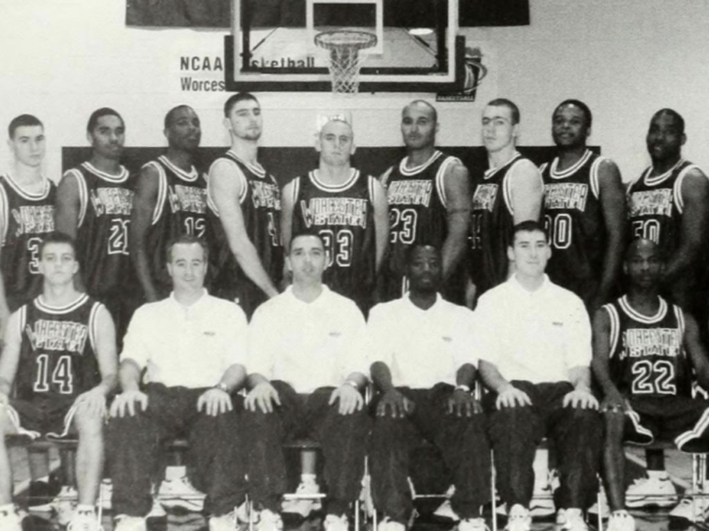 A basketball team photo commemorating their anniversary, featuring ten players standing in the back row and five coaches or team staff sitting in the front row. The background includes a basketball hoop and an NCAA Volleyball banner.