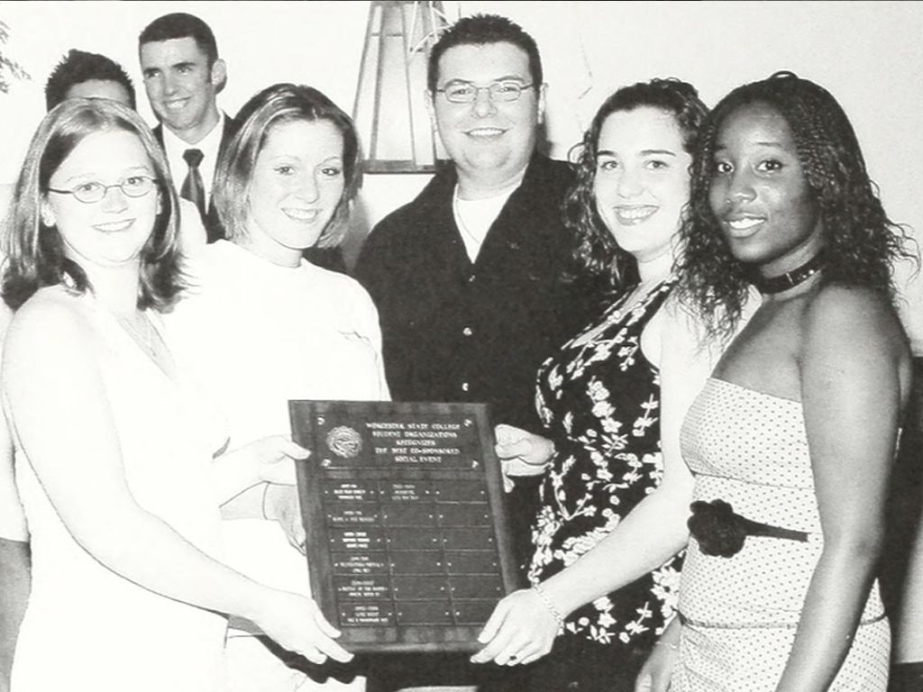 A group of five young adults, three women and two men, pose indoors holding a large rectangular plaque celebrating their anniversary. Two women hold the plaque at the front, while the others stand closely in the background.