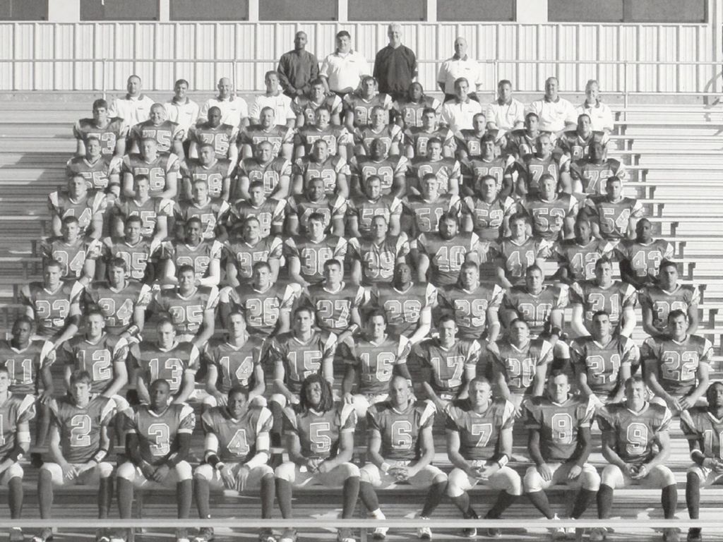 A black-and-white group photo of a football team in uniforms, seated and standing on bleachers. Coaches and team members are arranged in rows, facing the camera, capturing a moment that commemorates their anniversary season together.