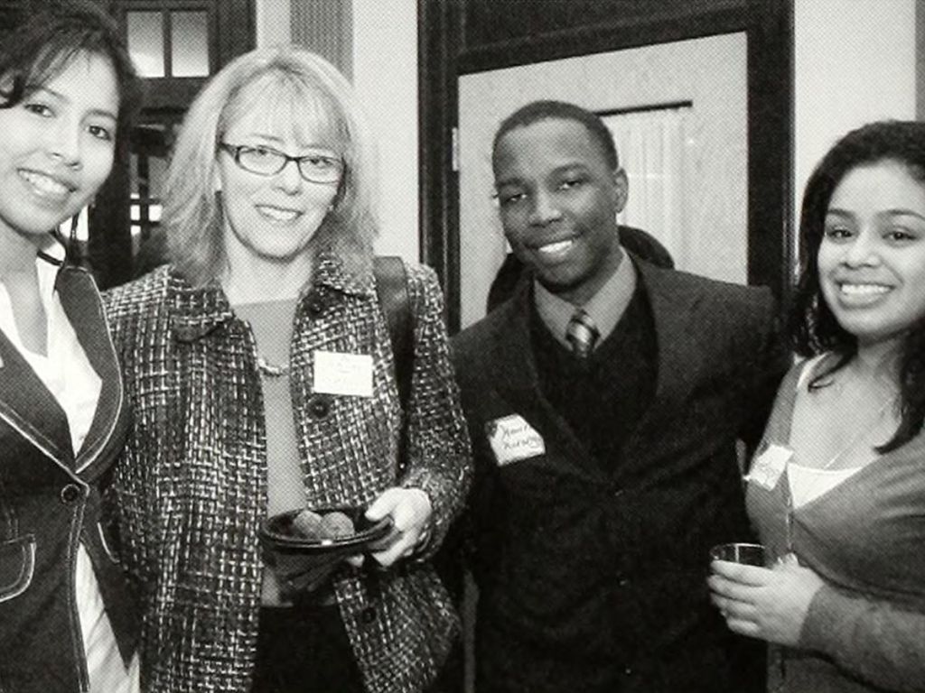 Four people are standing close together at a formal event, smiling at the camera. Two of them are holding drinks and visible name tags are worn by three individuals. It appears to be an anniversary celebration.