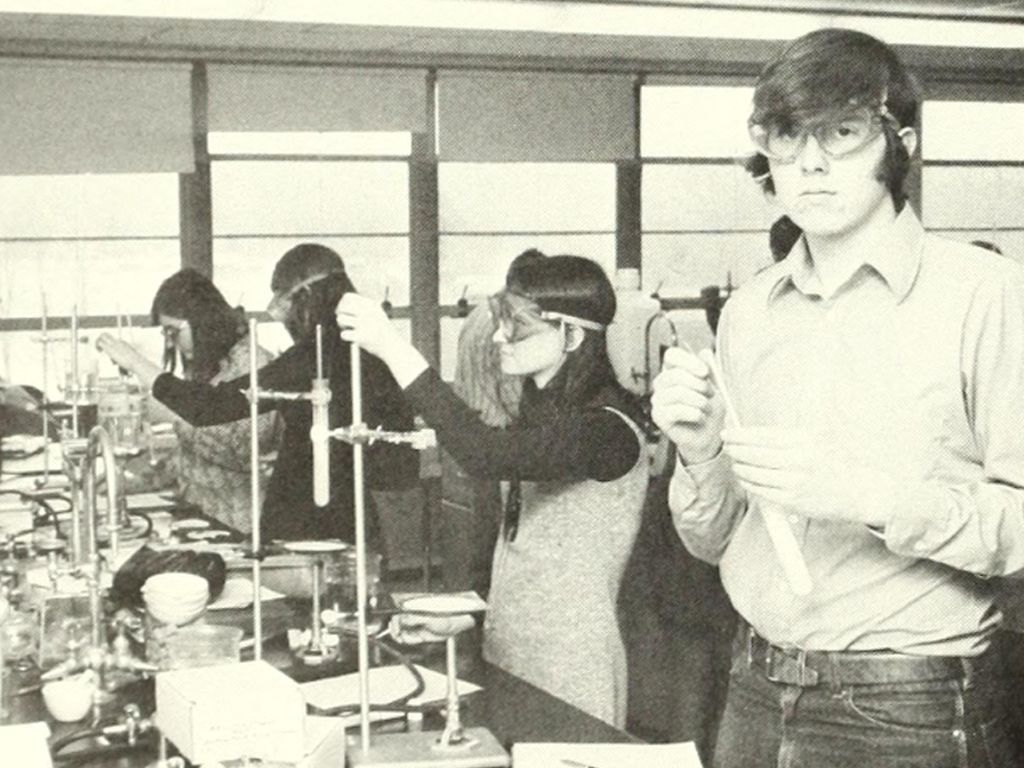 A group of students in a science lab is celebrating their school’s anniversary by conducting experiments. They are wearing safety goggles and handling test tubes and other laboratory equipment.