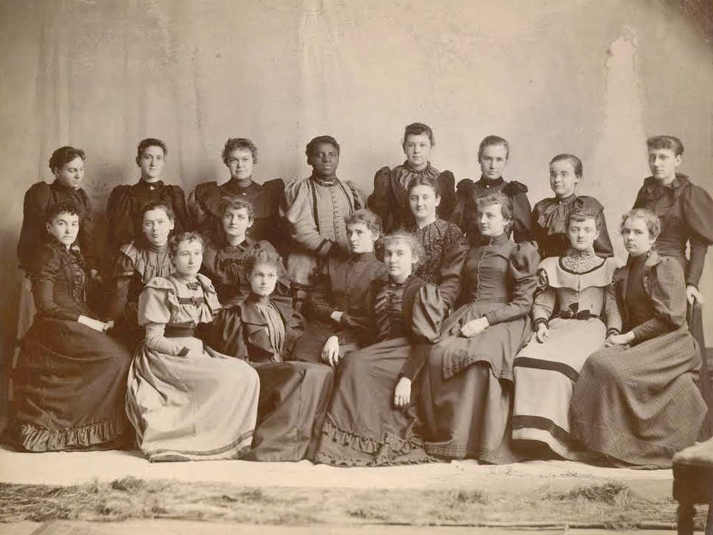 A sepia-toned photograph of 17 women from the late 19th or early 20th century, posed in two rows—most seated and a few standing—all wearing formal dresses of the period. The image was taken to commemorate an anniversary, capturing a moment steeped in history and elegance.