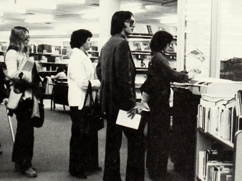 People standing in line at a library counter, some holding books and papers, with bookshelves visible in the background, celebrating the library's anniversary.