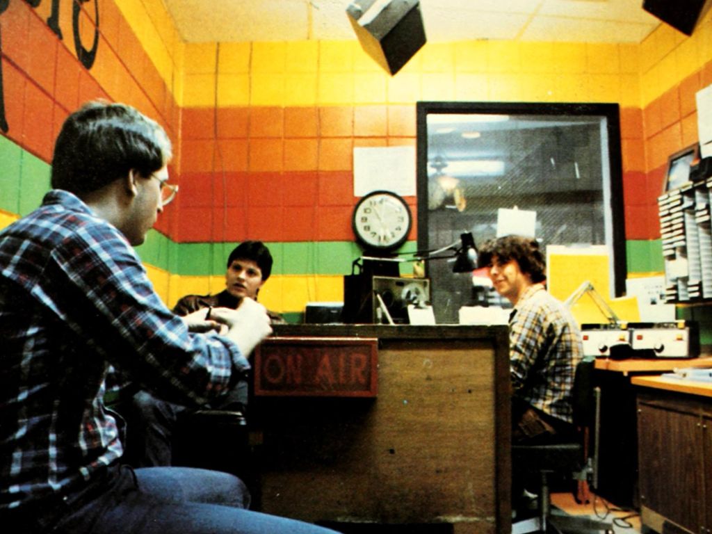 Three people are sitting in a brightly colored radio station studio. One person is wearing headphones, another is talking, and the third is smiling. An "ON AIR" sign is visible on a desk, adding to the festive atmosphere of their anniversary broadcast.