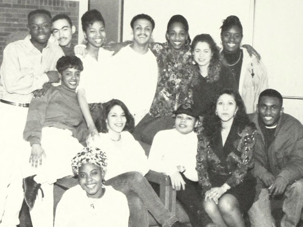 A group of thirteen people pose together indoors, celebrating an anniversary. Some are seated while others stand behind, all smiling warmly.