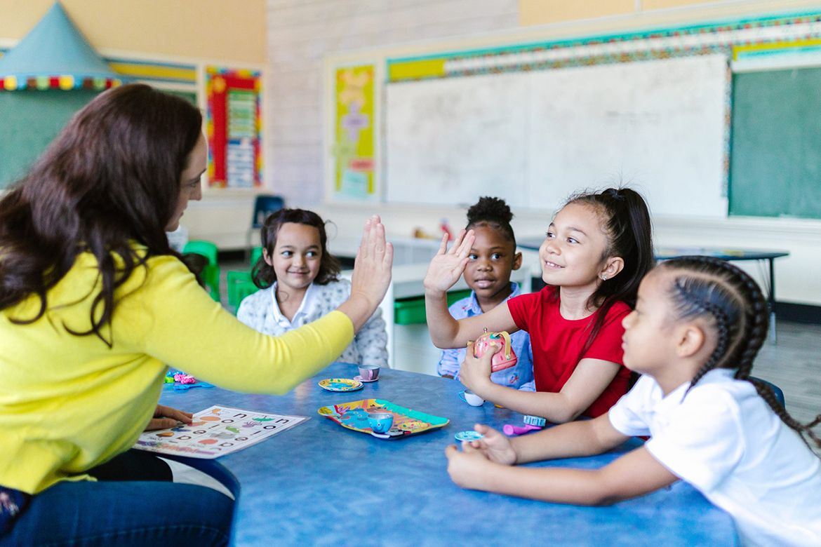 A teacher and four young students sit at a table in a classroom. One student gives the teacher a high-five while the others observe and smile, reflecting their joy in learning humanities. Learning materials and toys are on the table, fostering an engaging educational environment.