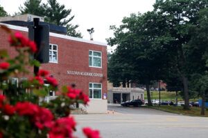 A brick building labeled "Sullivan Academic Center" is surrounded by trees and a parking area with several cars. Red flowers are visible in the foreground.