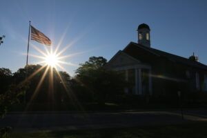 A building with a white columned entrance and a steeple is silhouetted against the sun, which is shining brightly. An American flag on a pole stands to the left of the building.