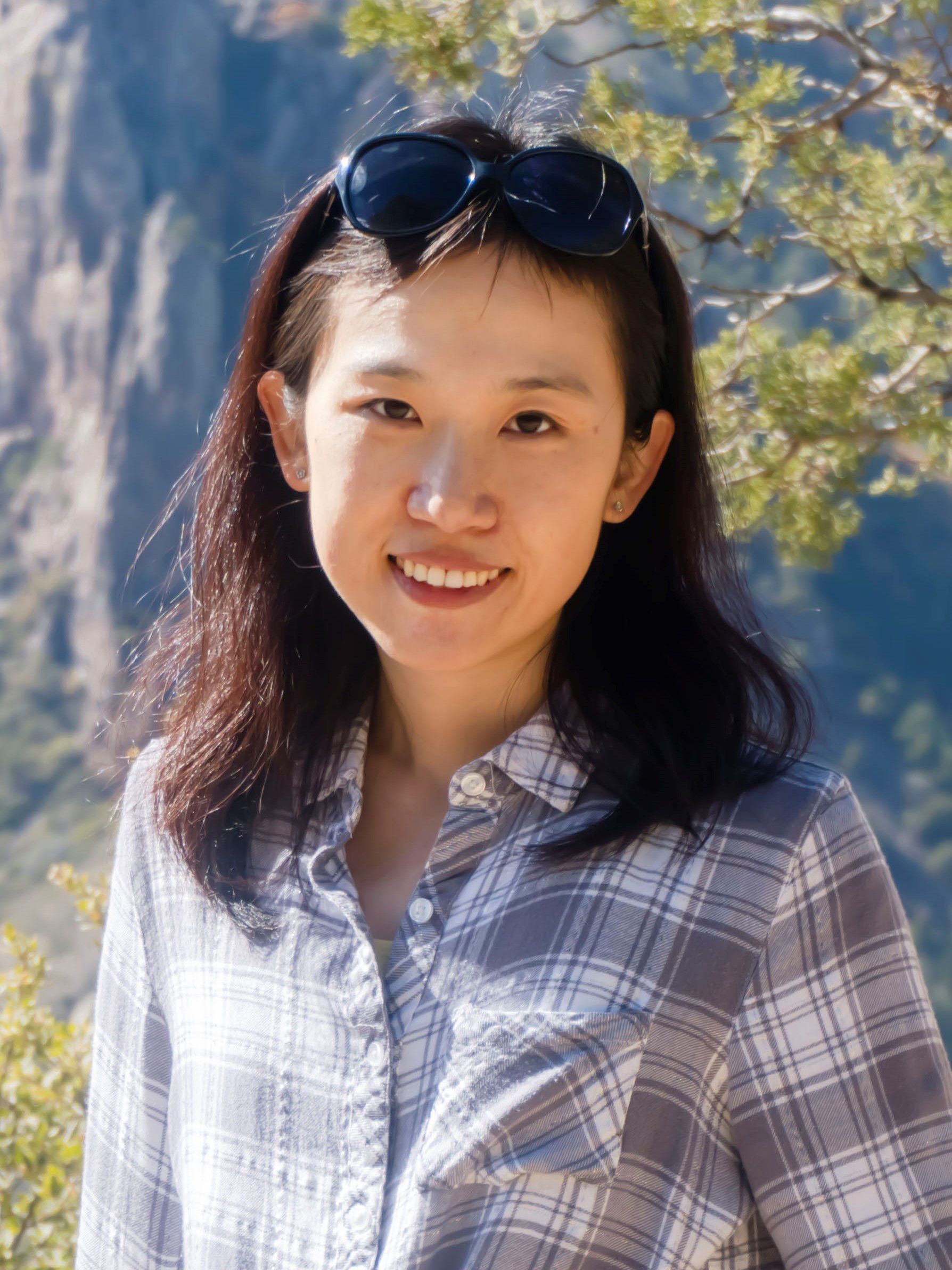 A woman with sunglasses on her head smiles at the camera, wearing a plaid shirt. Trees and rocks are visible in the background.