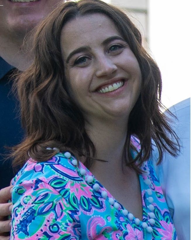 A woman with wavy brown hair smiles at the camera. She is wearing a colorful dress with blue, pink, and green patterns.