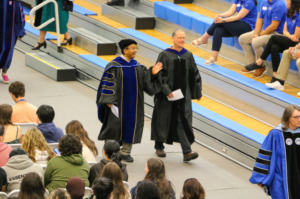 Two people in academic regalia walk through a gymnasium while a seated audience watches. One person waves to the crowd; the other person walks beside them.