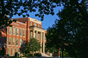 A red-brick academic building with large white columns and an inscription "School of Education Building" on its facade, surrounded by trees under a clear blue sky.