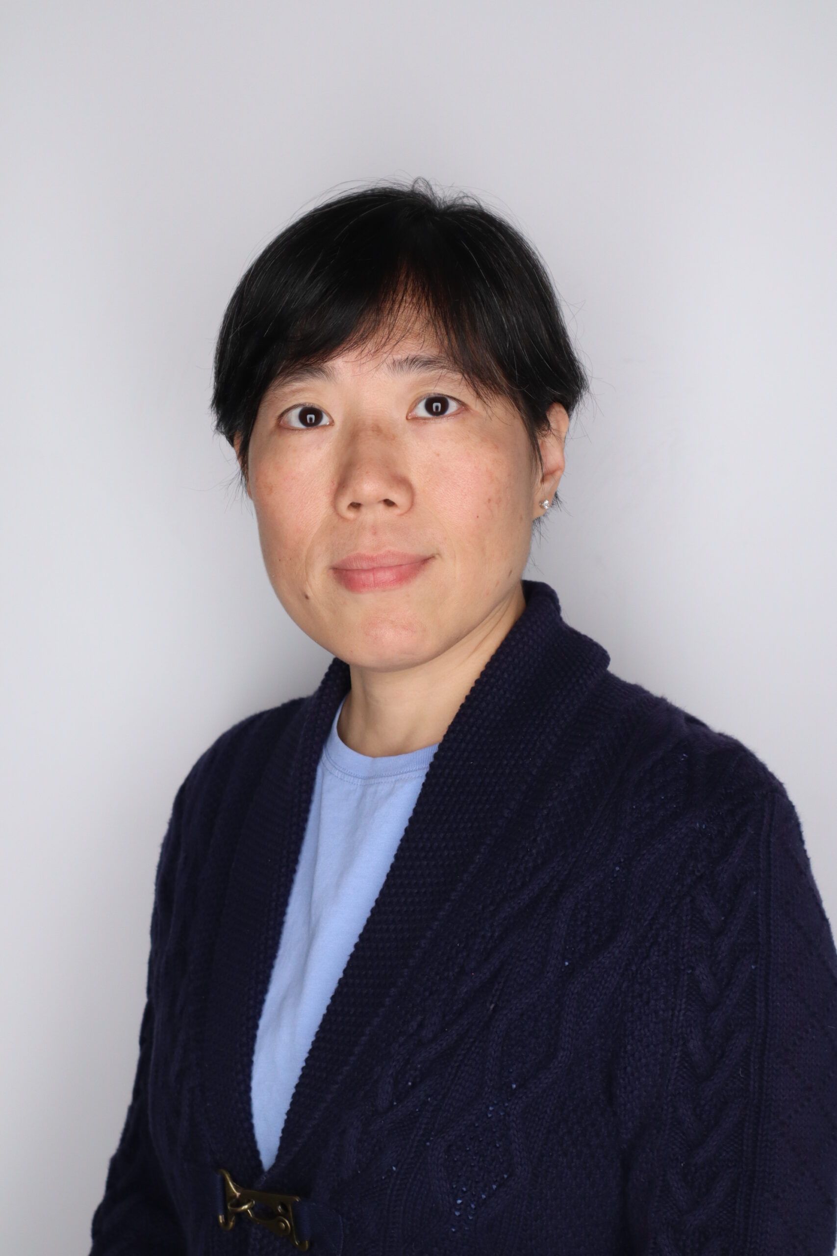 A woman with short black hair, wearing a navy blue cardigan over a light blue top, stands against a plain white background.