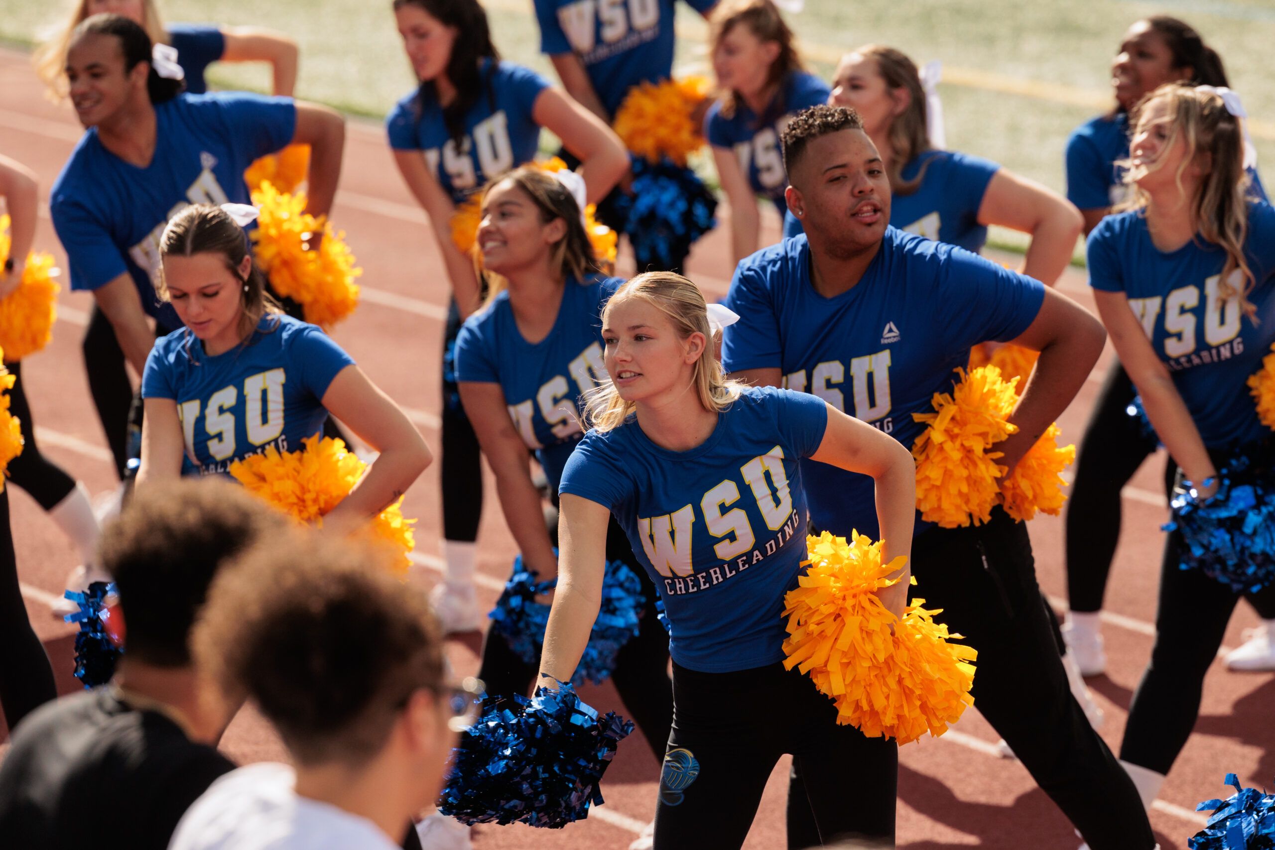 A cheerleading squad in blue "Worcester State University Cheerleading" shirts, holding yellow and blue pom-poms, performing a routine on a track field.