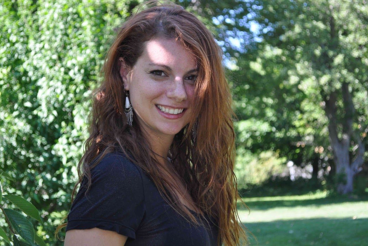 Smiling person with long, wavy hair stands outdoors near trees, wearing a black shirt and earrings.