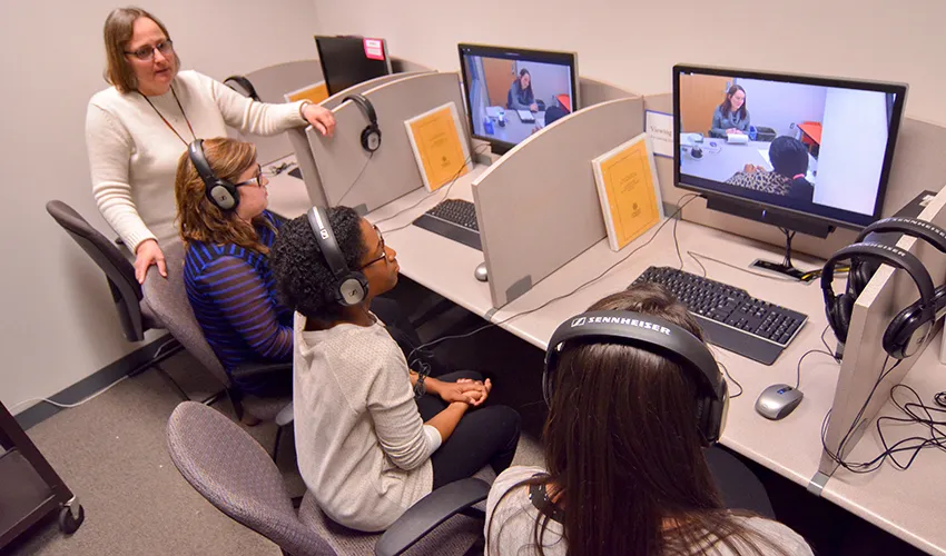 People wearing headphones sit at cubicles, engaging with business administration videos on computer monitors in a classroom setting, while an instructor observes.