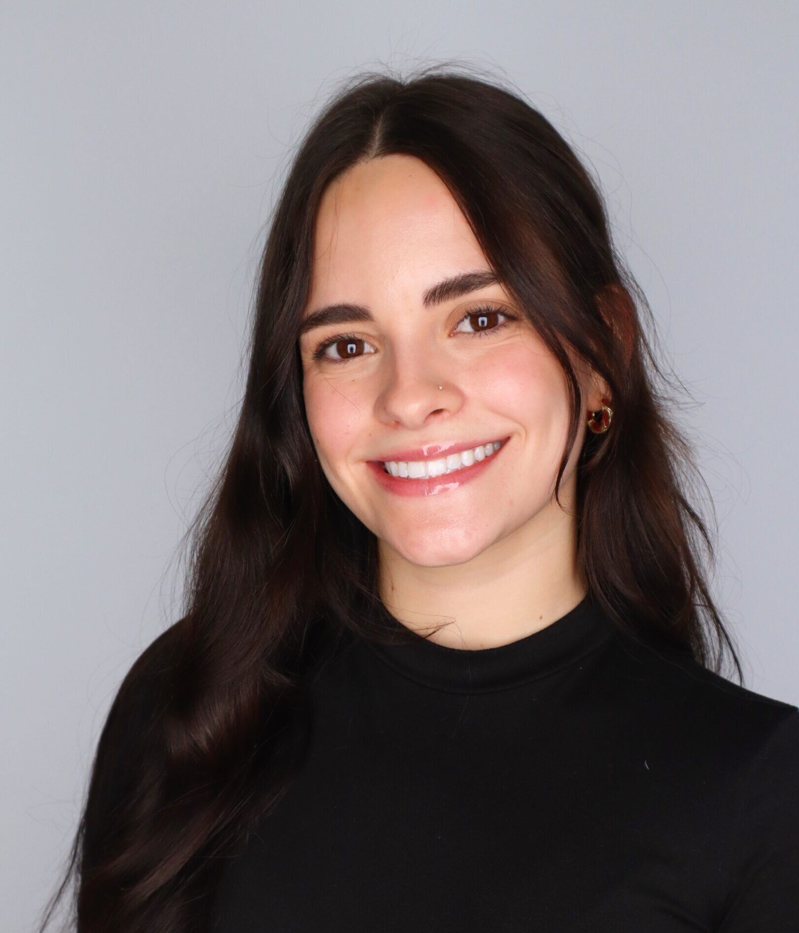 A woman with long dark hair wearing a black long-sleeve top smiles at the camera in front of a plain gray background.