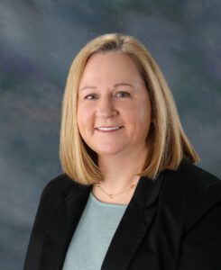 Portrait of a smiling person with shoulder-length blonde hair, wearing a black blazer and light green blouse against a dark background.