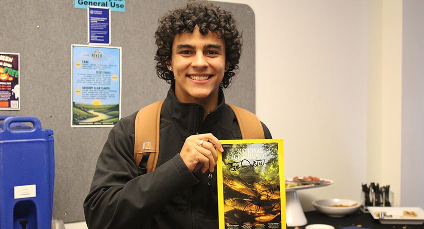 A person smiles warmly, holding a National Geographic magazine in a vibrant room adorned with Worcester State University posters. A table laden with refreshing treats adds to the inviting atmosphere.