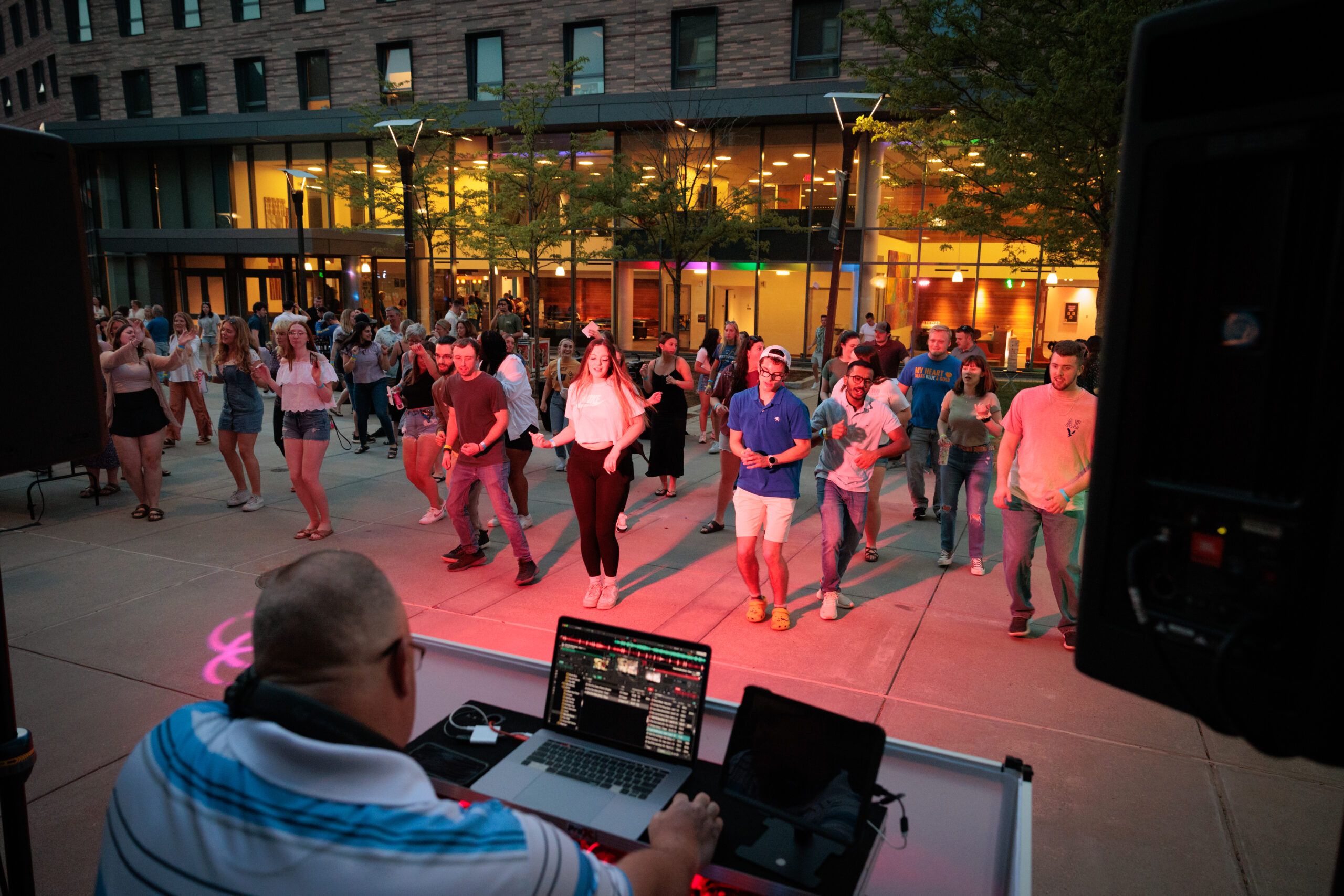 A group of people dancing outdoors on a patio in front of a DJ, with a building in the background.
