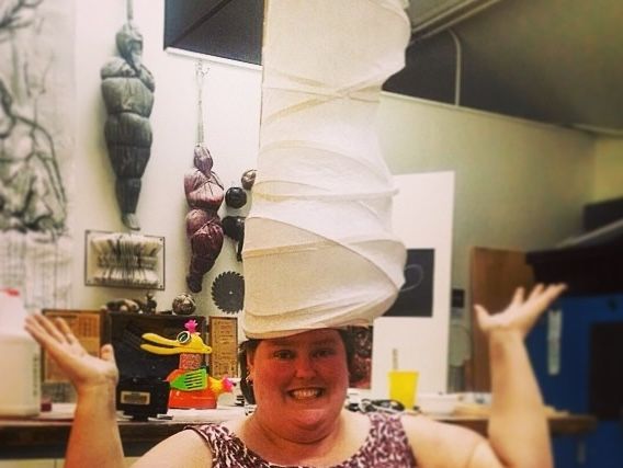 Person smiling with a large, sculptural hat in an art studio, surrounded by various artworks and materials.