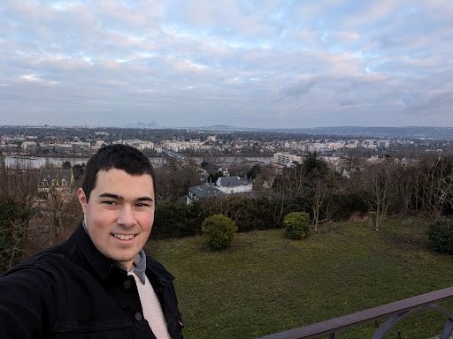 Person smiling for a selfie with a view of a cityscape and cloudy sky in the background.