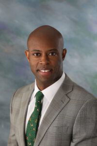 A man in a gray suit with a green tie stands against a cloudy blue backdrop, looking into the camera.
