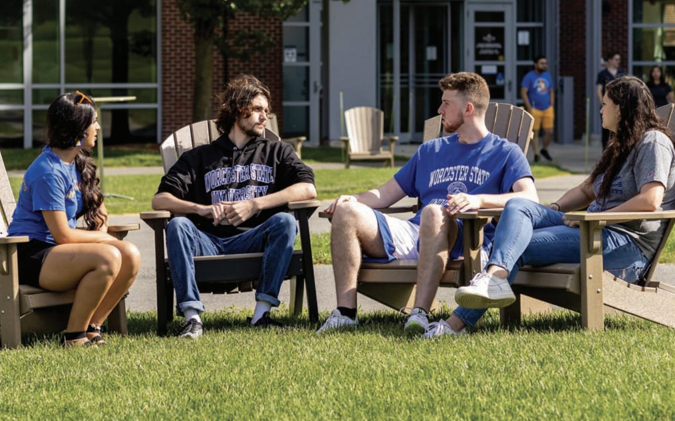 Four students sit in chairs on a grassy area outside a building, wearing casual attire. A banner at the bottom reads, "Make an Investment to Last Your Lifetime.
