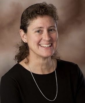 A woman with curly brown hair, wearing a black top and a silver necklace, smiles at the camera against a brown and beige background.