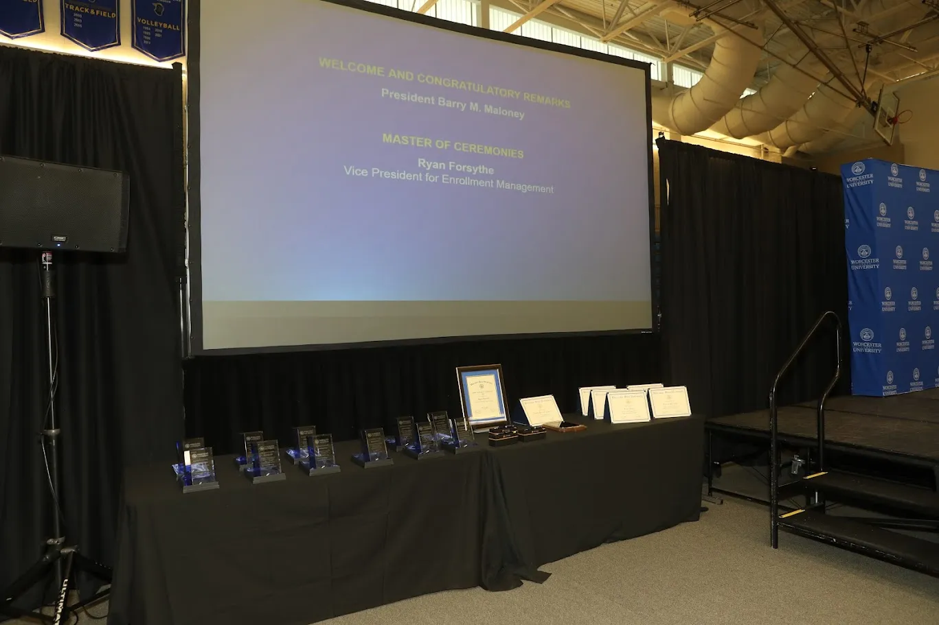 Stage setup for an awards ceremony with a projection screen displaying event details, a table of trophies and certificates, and a step leading to the stage.