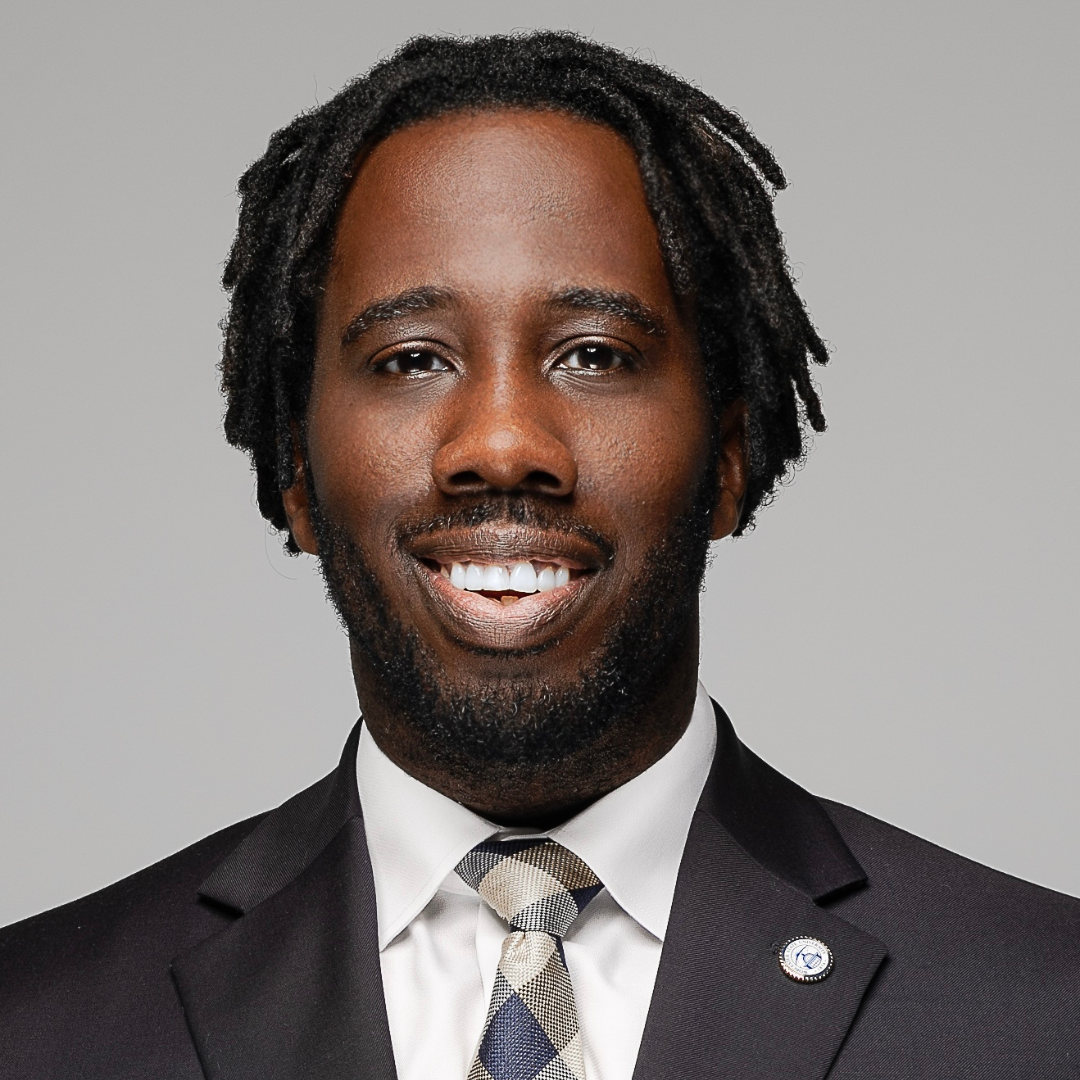 Smiling man with short dreadlocks wearing a dark suit, checkered tie, and a lapel pin, posed against a plain gray background.