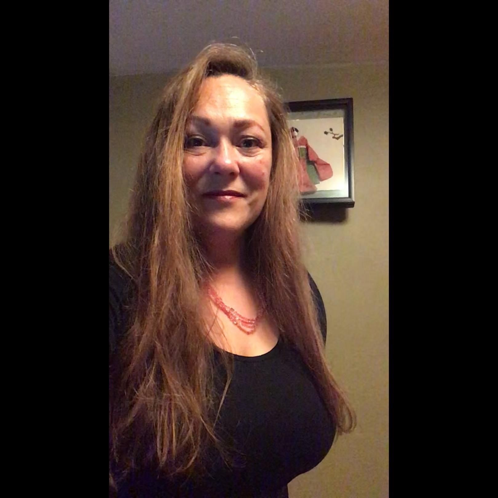 A woman with long brown hair wearing a black top and red necklace stands indoors with a framed picture on the wall behind her.