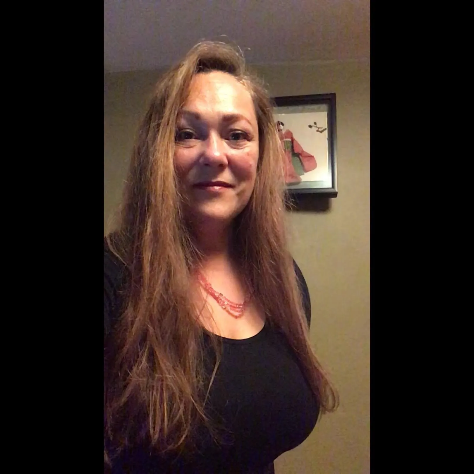 A woman with long brown hair wearing a black top and red necklace stands indoors with a framed picture on the wall behind her.