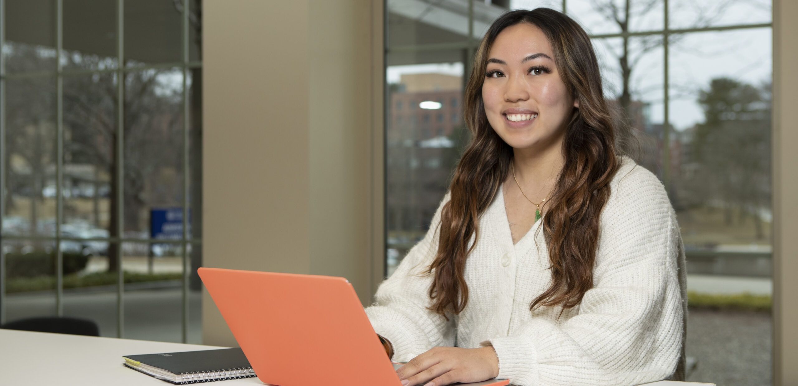 A woman sits at a desk with an orange laptop, smiling at the camera. A notebook and pen are on the desk. Large windows show an outdoor scene in the background.