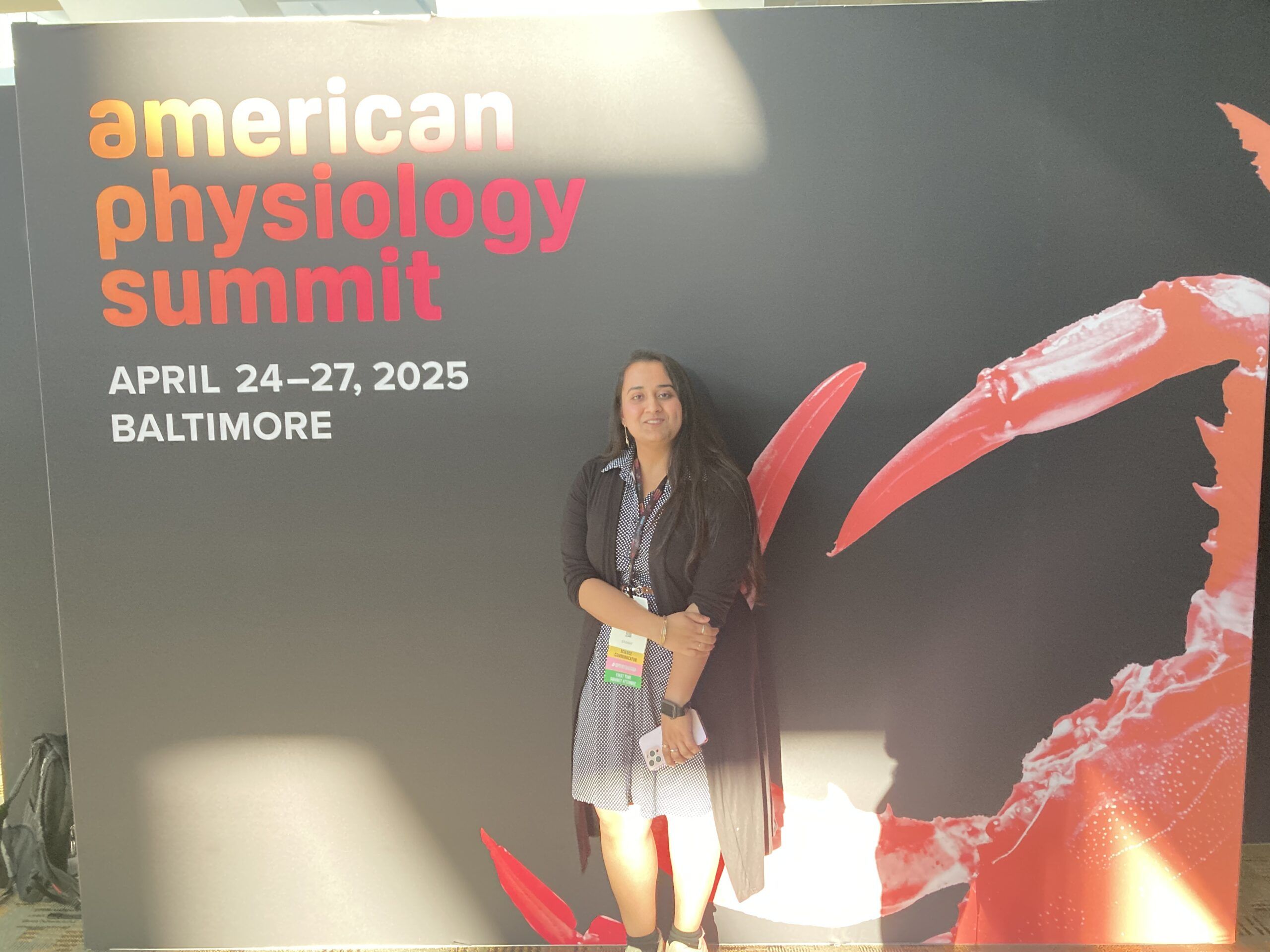A person stands in front of a sign that reads "American Physiology Summit, April 24-27, 2025, Baltimore" with a large red crab claw graphic in the background.