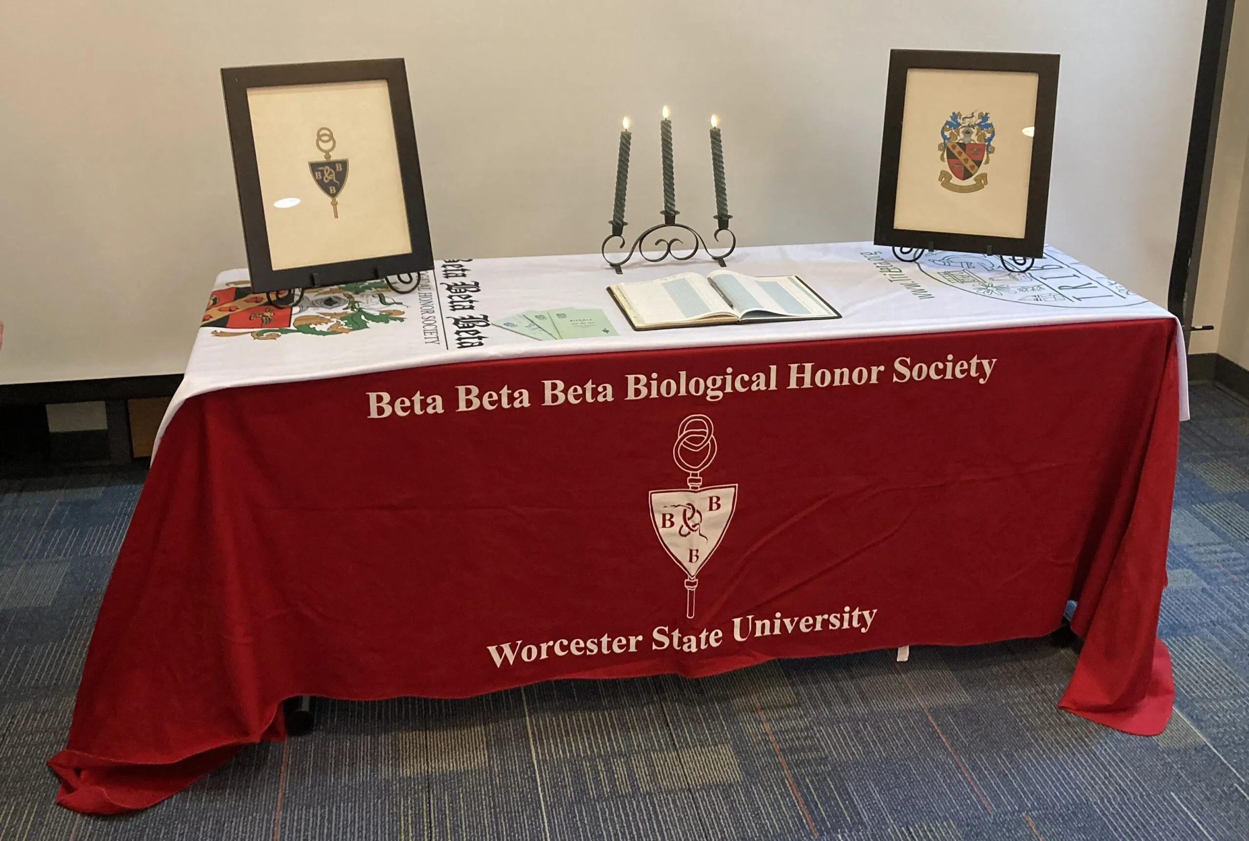 A table with a red cloth labeled "Beta Beta Beta Biological Honor Society, Worcester State University," with framed images, candles, and an open book displayed on top.