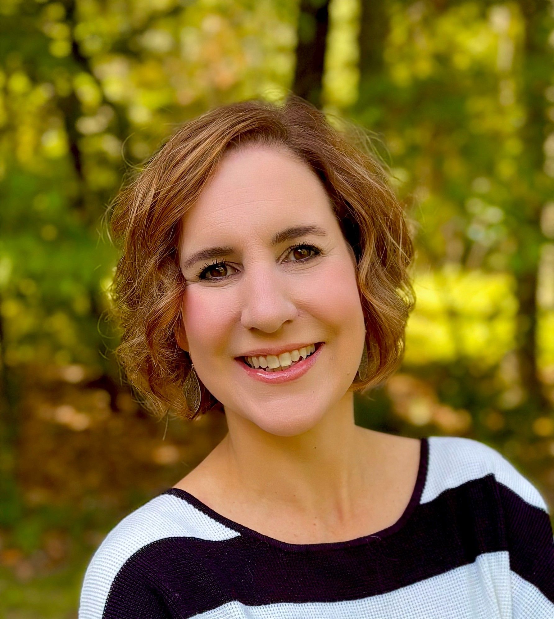 A woman with short, wavy brown hair smiles outdoors. She is wearing a white and black striped top with a blurred green foliage background.