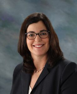 Woman with dark hair and glasses wearing a dark blazer and light blouse, smiling at the camera, in front of a gray and blue mottled background.