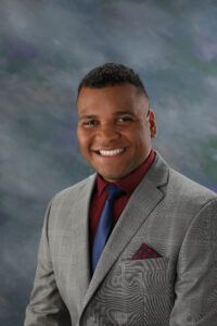 A man wearing a gray suit, burgundy shirt, and blue tie smiles at the camera against a marbled blue and gray studio backdrop.