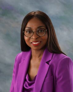 A woman with long dark hair, wearing round glasses and a purple blazer, poses in front of a softly blurred, neutral-toned background.