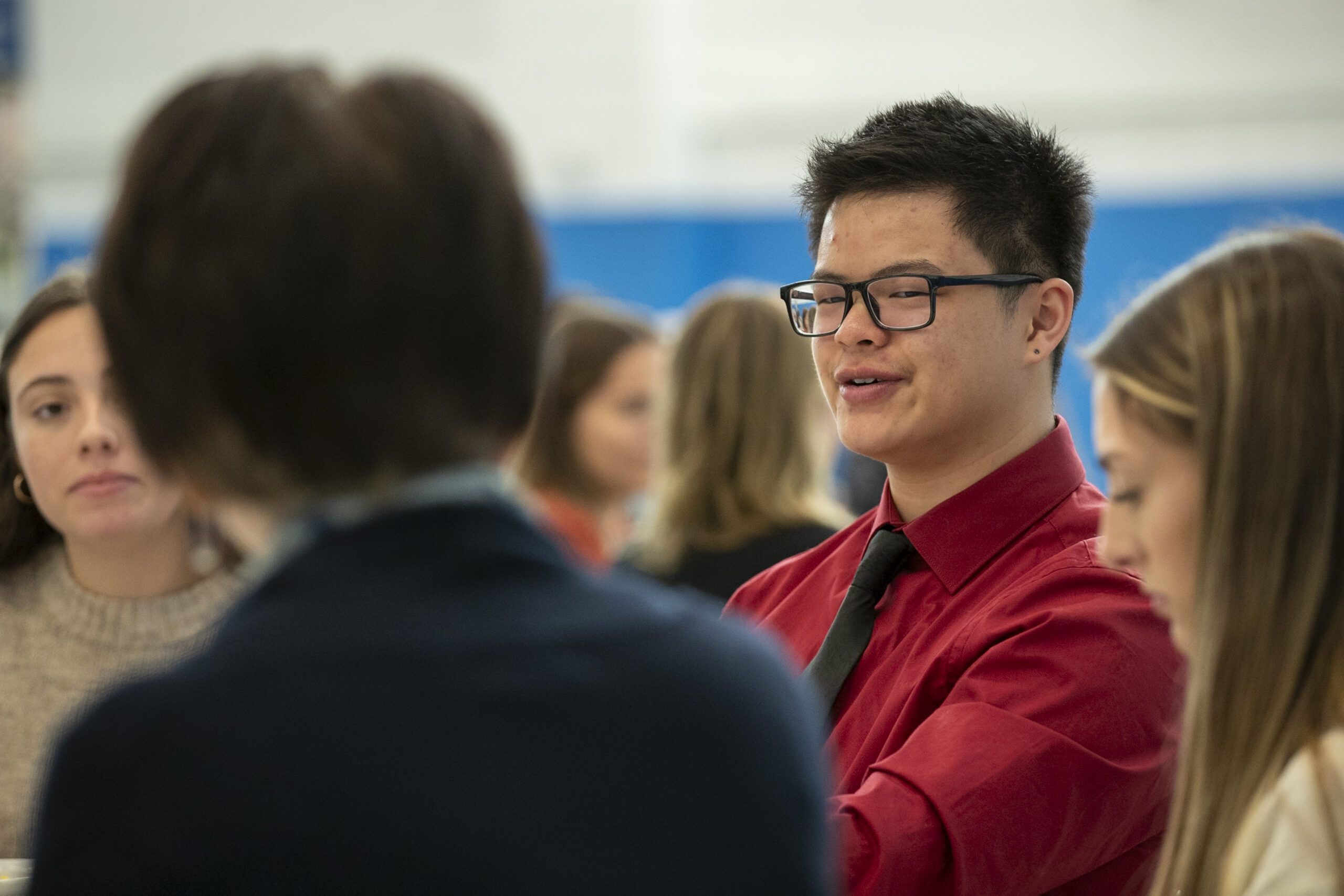 A group of people sit together at a table, with one person in a red shirt and glasses speaking.