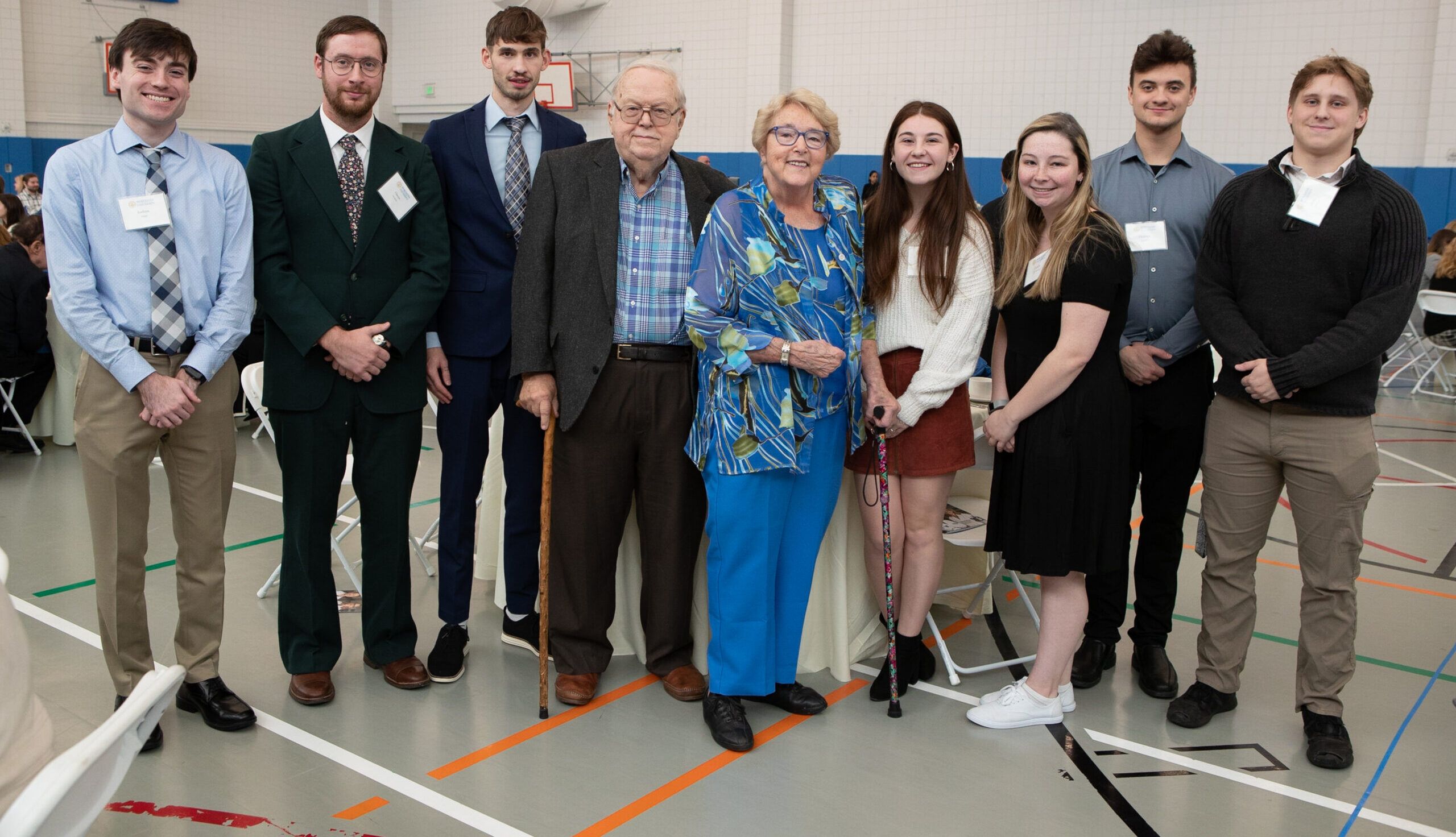 A group of eight people, including six young adults and two older adults, posing together in a gymnasium during what appears to be a formal or semi-formal event.