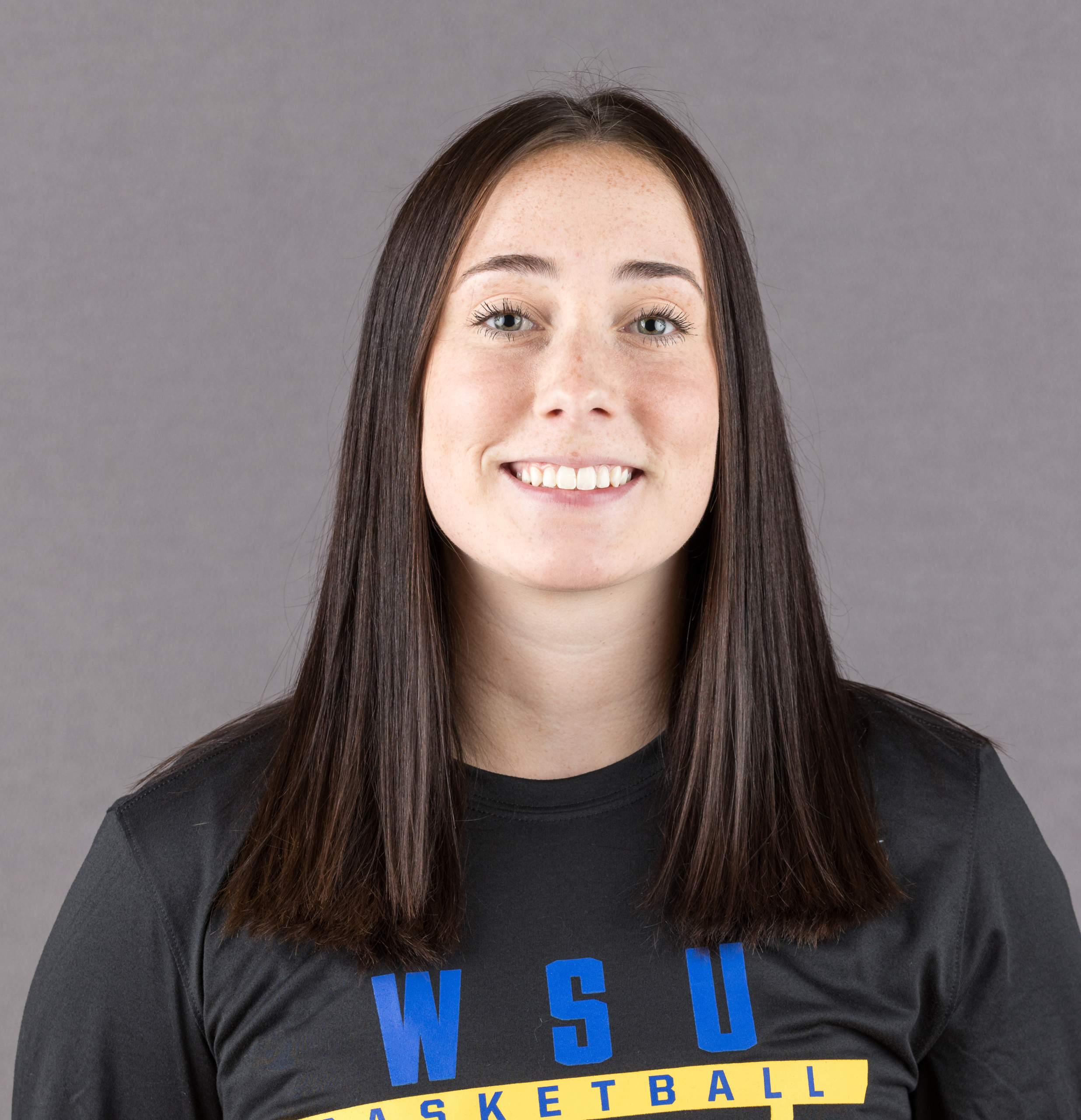 A young woman with straight brown hair smiles at the camera, wearing a black T-shirt with blue and yellow "WSU BASKETBALL" text, against a plain gray background.