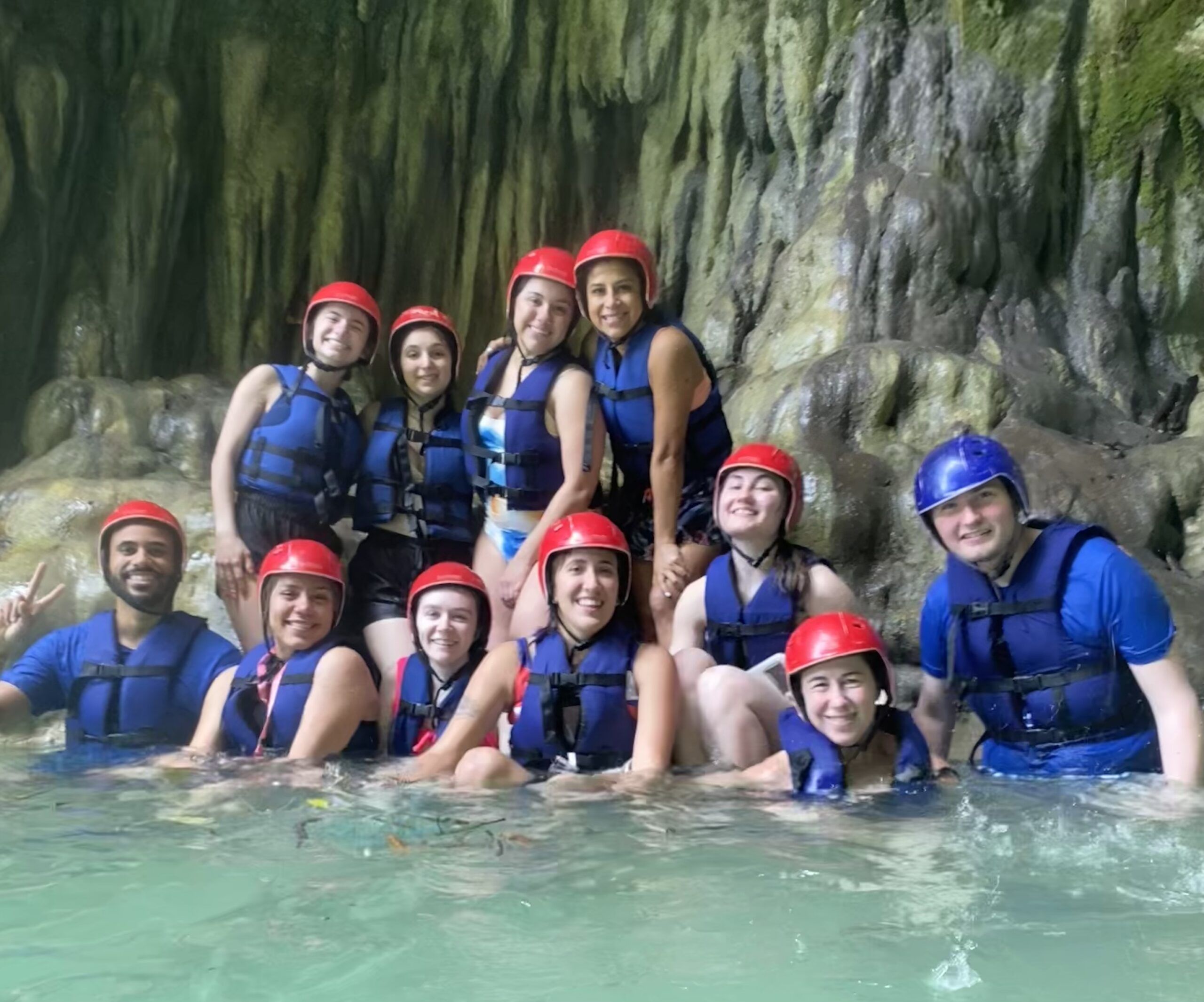 A group of people wearing helmets and life jackets pose together in shallow water inside a rocky cave.