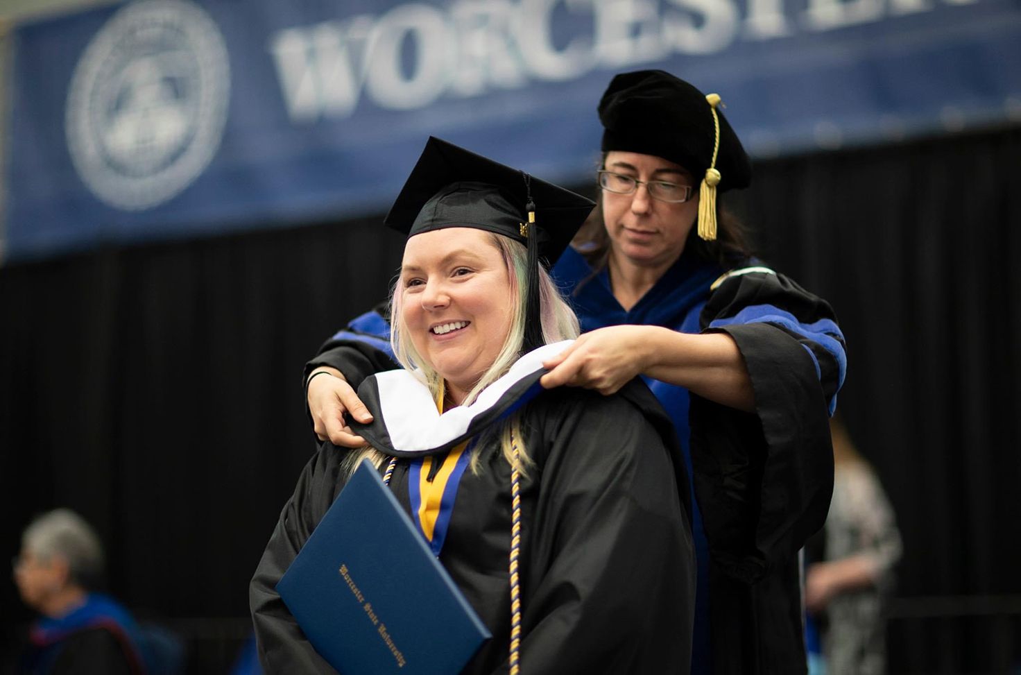 A graduate is hooded by a faculty member during a commencement ceremony, holding a diploma folder, with a "Worcester" banner visible in the background.