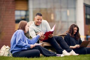 Three students sit on grass outside a building; two discuss notes from a folder, and one works on a laptop.