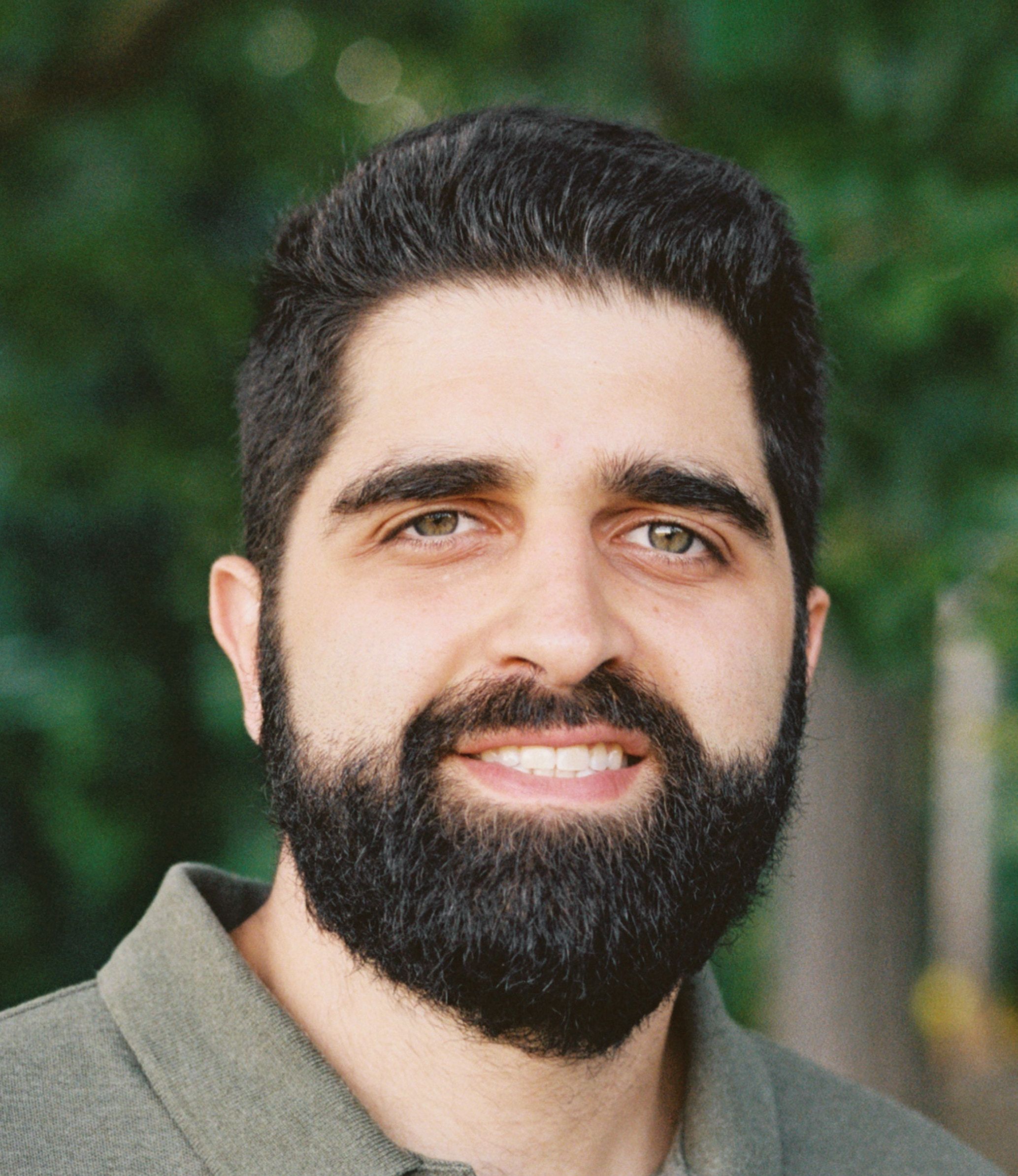 A man with dark hair and a full beard is smiling outdoors, wearing a green collared shirt. Green foliage is visible in the background.