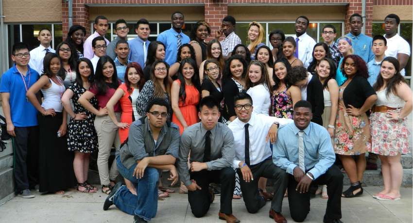 A large group of young adults poses together outdoors in front of a building at Worcester State University, dressed in a mix of business casual and semi-formal attire.