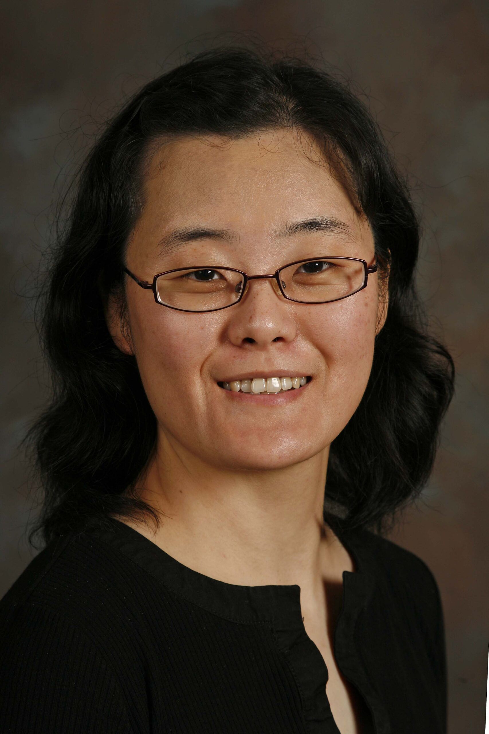 A woman with medium-length black hair and glasses is smiling at the camera against a dark, blurred background. She is wearing a black top.