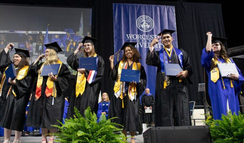 Six graduates in caps and gowns celebrate on stage with diplomas at Worcester State University's ceremony, a proud moment made possible for many through tuition support and financial aid, with the university banner displayed in the background.